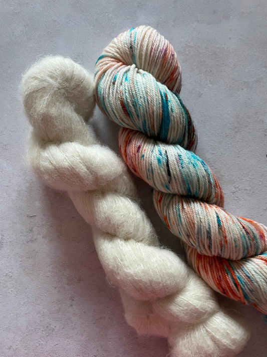 A skein of fluffy suri alpaca and silk yarn and a skein of double knitting weight with blue and orange speckles